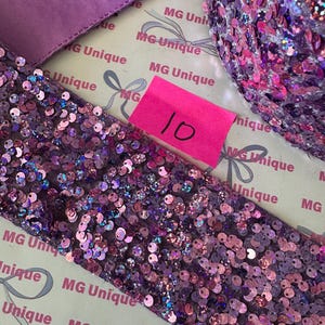 Op de afbeelding: Close-up van paars paillettenlint en stof. Het lint is bedekt met kleine, iriserende pailletten in paarse en roze tinten. Een roze plakbriefje met het nummer 10 is zichtbaar. De achtergrond bevat gedrukte tekst.
