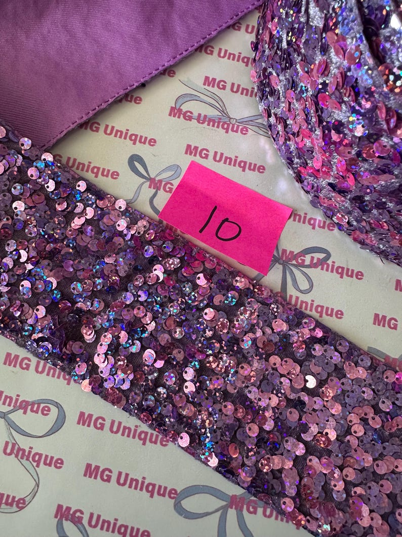 Op de afbeelding: Close-up van paars paillettenlint en stof. Het lint is bedekt met kleine, iriserende pailletten in paarse en roze tinten. Een roze plakbriefje met het nummer 10 is zichtbaar. De achtergrond bevat gedrukte tekst.