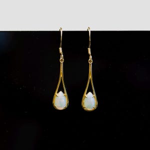 May include: A pair of gold-tone drop earrings featuring pear-shaped opal stones. The earrings have a slender, open frame design and a hook fastening. The opals are a milky white colour.