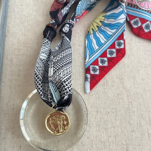 May include: A clear, circular glass pendant with a gold-colored medallion featuring a relief design, suspended from a patterned silk scarf. The scarf displays a variety of colors and geometric patterns, including red, blue, and black.