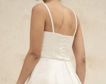 May include: A white wedding dress with a fitted bodice and spaghetti straps. The dress has a pleated skirt that flares out from the waist. The back of the dress features a row of buttons. The model is wearing pearl earrings.