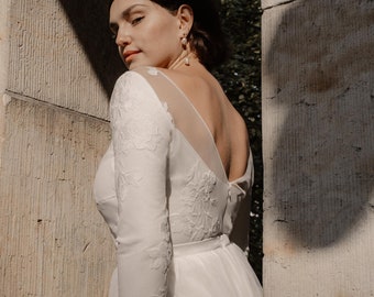 May include: A white wedding dress with long sleeves and a low back. The dress features delicate floral embroidery and a sheer panel on the back.