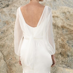 Könnte beinhalten: Ein weißes Hochzeitskleid mit tiefem, offenem Rücken und langen, transparenten Ärmeln. Das Kleid hat eine Schleife an der Taille und einen fließenden Rock. Der Stoff scheint ein weiches, leichtes Material zu sein.