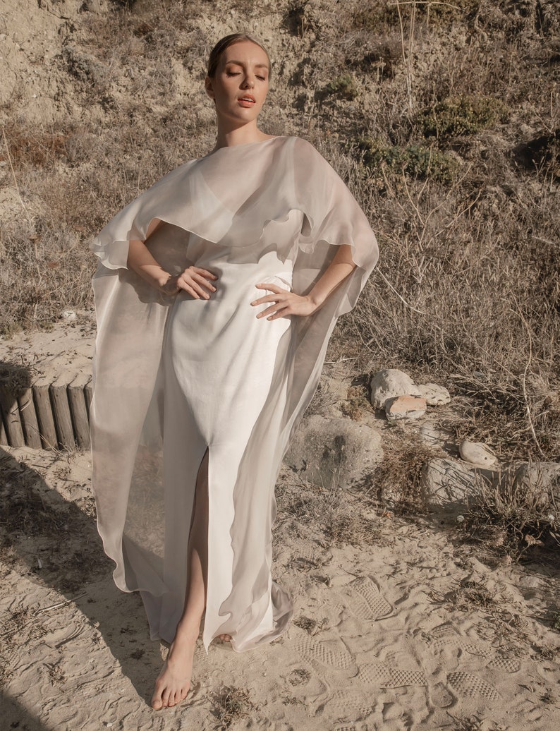 May include: A flowing, white gown with a high slit, paired with a sheer, cape-like overlay. The cape is a light, neutral color, creating a layered effect. The outfit is worn outdoors on a sandy surface, suggesting a beach or desert setting.
