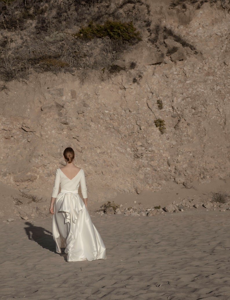 May include: A woman wearing a white satin wedding dress with a long train and a bow at the waist. She is walking on a sandy beach.