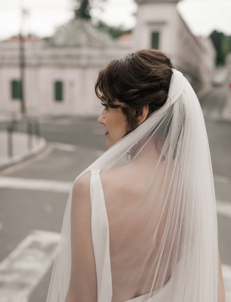 Puede incluir: Una novia con un vestido de novia blanco y un velo largo y fluido. El velo est&aacute; hecho de una tela transparente y est&aacute; drapeado sobre la cabeza y los hombros de la novia. La novia est&aacute; de pie frente a un edificio con una fachada blanca.