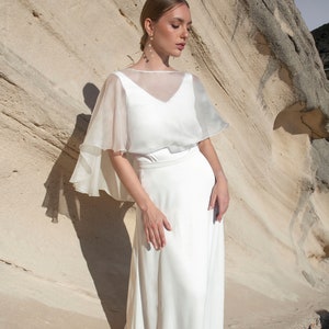 May include: A white bridal gown with a V-neck bodice and a flowing skirt. A sheer, cape-like overlay drapes over the shoulders. The model is standing in front of a light-colored rock formation.