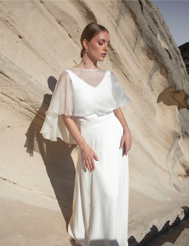 May include: A white bridal gown with a V-neck bodice and a flowing skirt. A sheer, cape-like overlay drapes over the shoulders. The model is standing in front of a light-colored rock formation.