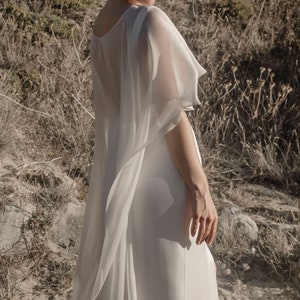 May include: A white, flowing dress with a sheer, cape-like overlay. The dress has a simple, elegant design, with a high neckline and a long, flowing skirt. The sheer overlay adds a touch of drama and sophistication. The model is standing outdoors in a natural setting.