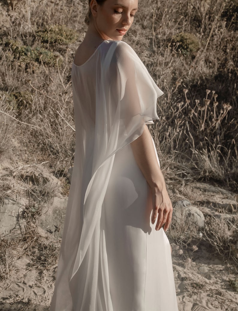 May include: A white, flowing dress with a sheer, cape-like overlay. The dress has a simple, elegant design, with a high neckline and a long, flowing skirt. The sheer overlay adds a touch of drama and sophistication. The model is standing outdoors in a natural setting.