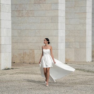 May include: A woman wearing a white strapless dress with a long train. The dress has a fitted bodice and a flowing skirt. The woman is walking on a cobblestone path in front of a white brick wall.