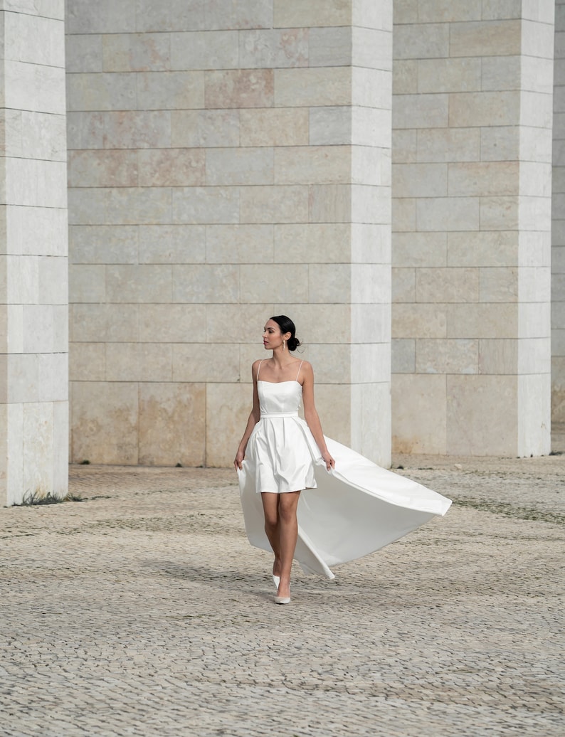 May include: A woman wearing a white strapless dress with a long train. The dress has a fitted bodice and a flowing skirt. The woman is walking on a cobblestone path in front of a white brick wall.
