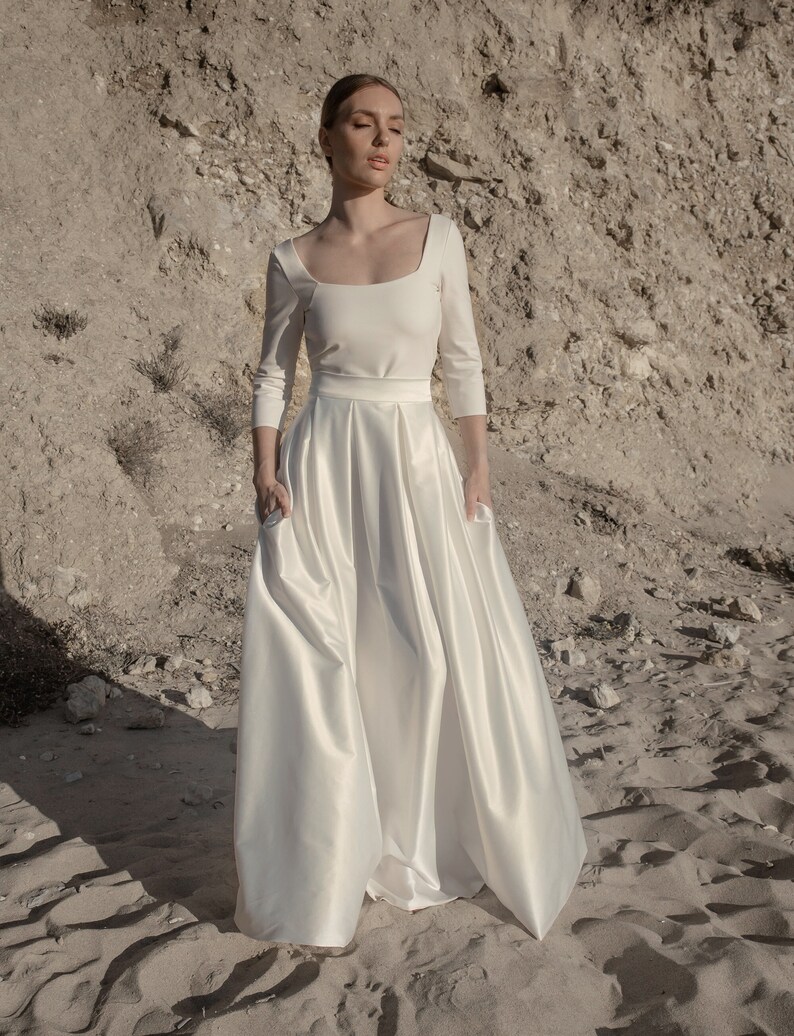 May include: A white satin wedding dress with a square neckline and three-quarter sleeves. The skirt is full and flowing, with a high waistline. The dress is photographed against a backdrop of sand and rock.