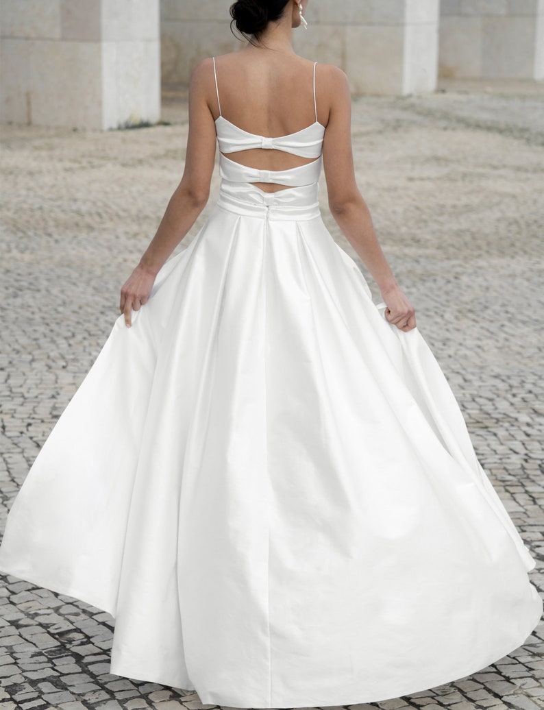 May include: A white satin wedding dress with a low back and a bow detail. The dress has a full skirt and a fitted bodice.
