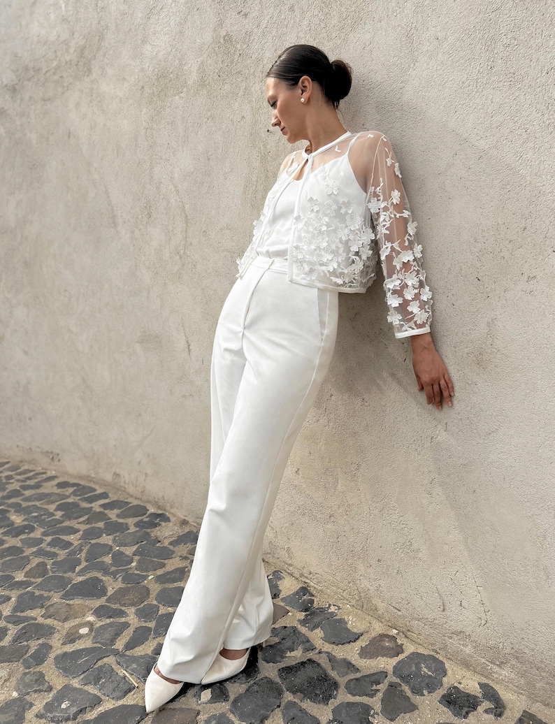 May include: A white sheer jacket with floral embroidery, worn over a white top and white trousers. The jacket has a V-neck and long sleeves. The model is standing against a white wall.