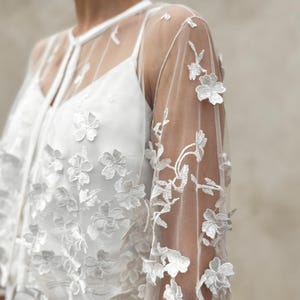 May include: White sheer top with floral embroidery. The top has long sleeves and a round neckline. The embroidery is delicate and intricate, and the top is perfect for a special occasion.
