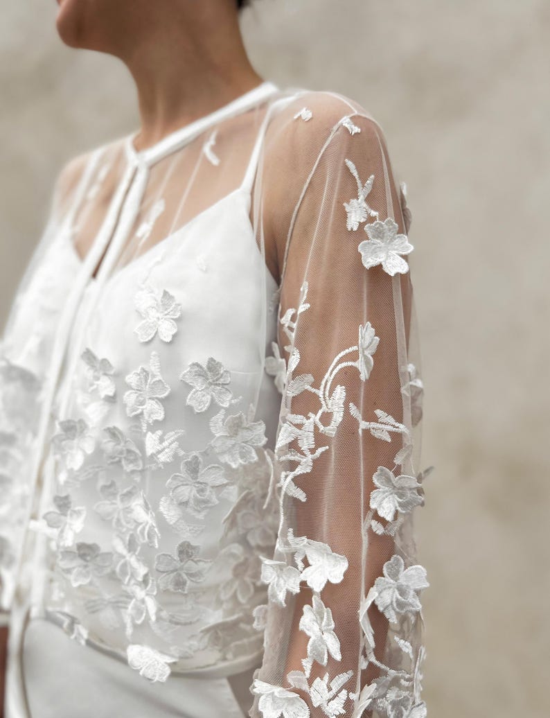 May include: White sheer top with floral embroidery. The top has long sleeves and a round neckline. The embroidery is delicate and intricate, and the top is perfect for a special occasion.