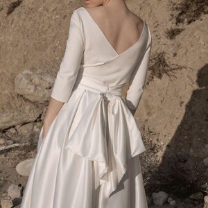 May include: A white satin wedding dress with a deep v-neckline and a bow at the back. The dress has a fitted bodice and a full skirt. The dress is shown on a model standing in a desert landscape.