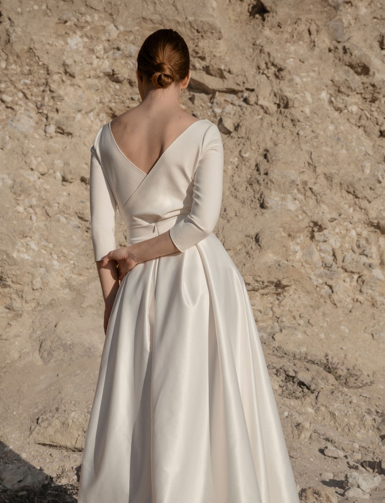 May include: A white satin wedding dress with a V-neckline and three-quarter sleeves. The dress has a fitted bodice and a flowing skirt.