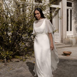 May include: A white wedding dress with a sheer overlay and flowing skirt. The dress is worn outdoors in front of a building with a stone facade and a green bush. The dress is a simple, elegant design.