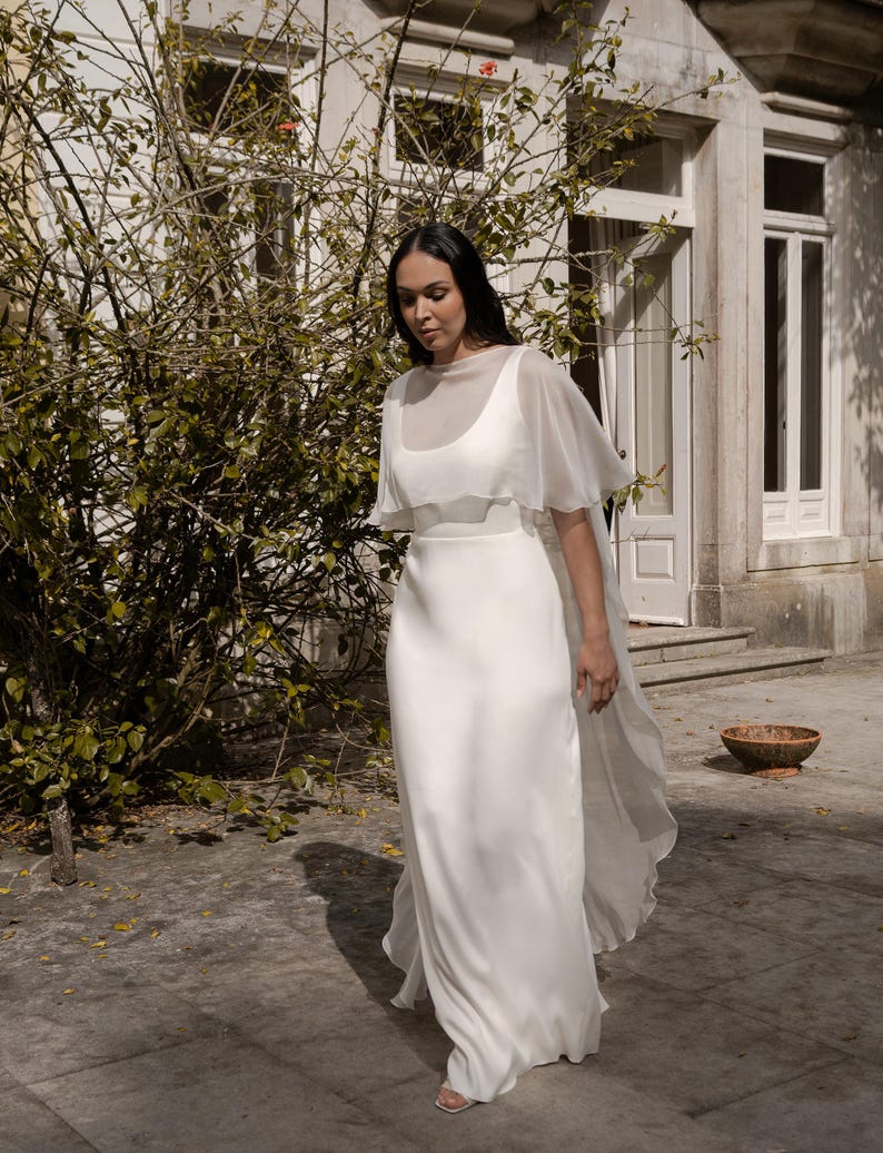 May include: A white wedding dress with a sheer overlay and flowing skirt. The dress is worn outdoors in front of a building with a stone facade and a green bush. The dress is a simple, elegant design.