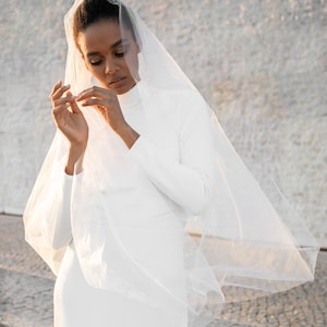 Peut inclure: Une femme portant une robe blanche et un voile transparent. Elle se tient devant un mur texturé.