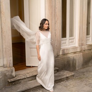 K&ouml;nnte beinhalten: Ein bodenlanges wei&szlig;es Kleid mit &auml;rmellosem Oberteil, V-Ausschnitt und einem transparenten, leichten &Uuml;berwurf, der sich an den Schultern zu einem flie&szlig;enden, capeartigen Element ausweitet und eine weich fallende Silhouette bis zum Boden bildet.