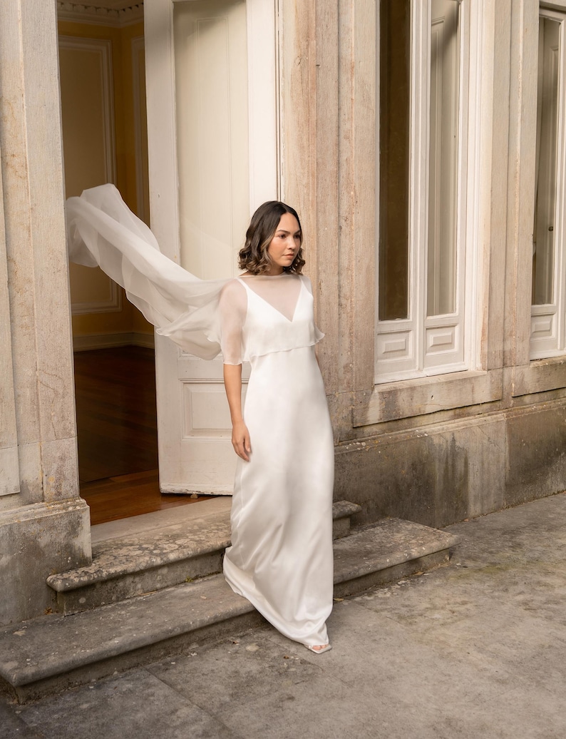 K&ouml;nnte beinhalten: Ein bodenlanges wei&szlig;es Kleid mit &auml;rmellosem Oberteil, V-Ausschnitt und einem transparenten, leichten &Uuml;berwurf, der sich an den Schultern zu einem flie&szlig;enden, capeartigen Element ausweitet und eine weich fallende Silhouette bis zum Boden bildet.