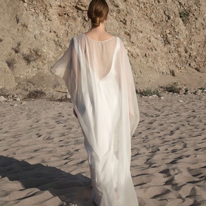 May include: A flowing white dress with a sheer cape, worn on a sandy beach. The dress has a low back and the cape drapes over the shoulders. The model is facing away from the camera, with the sun casting shadows on the sand.