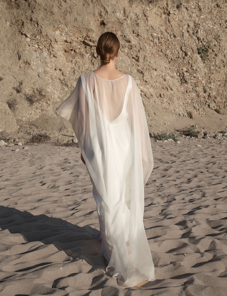 May include: A flowing white dress with a sheer cape, worn on a sandy beach. The dress has a low back and the cape drapes over the shoulders. The model is facing away from the camera, with the sun casting shadows on the sand.