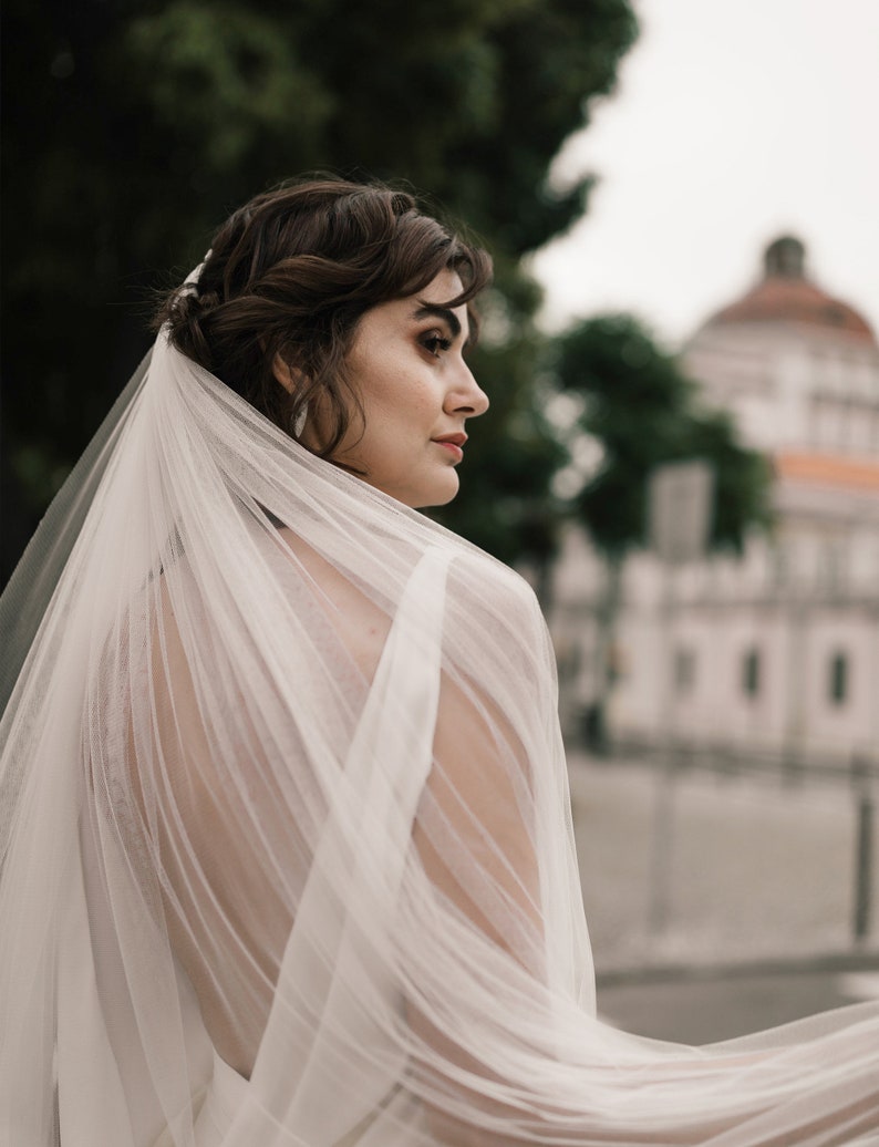 Puede incluir: Una novia con un velo blanco con un delicado patr&oacute;n de encaje. El velo fluye detr&aacute;s de ella mientras mira hacia un lado.