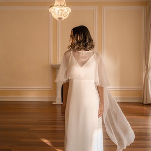 May include: A white wedding dress with a V-neckline and a flowing, sheer cape. The dress is floor-length and simple, with a minimalist design. The cape adds a touch of elegance and movement. The setting is a room with a chandelier.