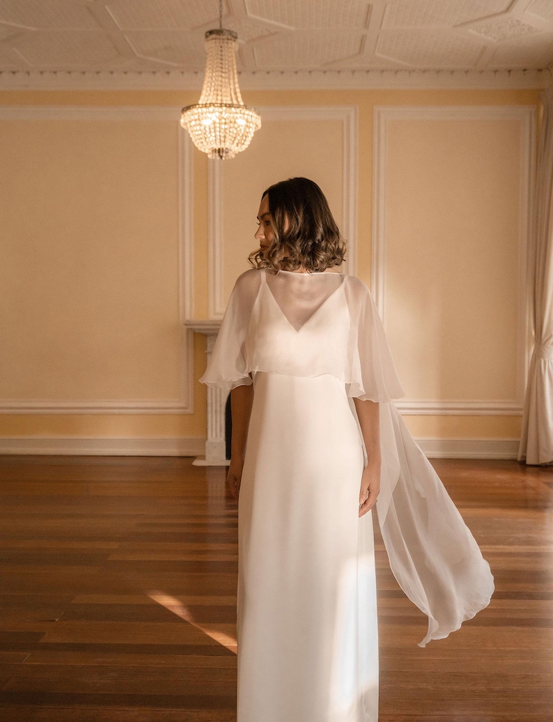May include: A white wedding dress with a V-neckline and a flowing, sheer cape. The dress is floor-length and simple, with a minimalist design. The cape adds a touch of elegance and movement. The setting is a room with a chandelier.