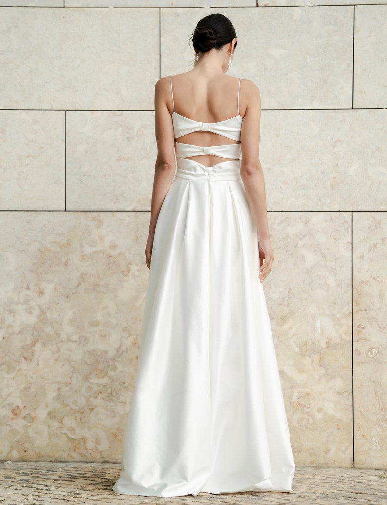 May include: A white satin dress with a low back and a bow detail. The dress has a fitted bodice and a flowing skirt.