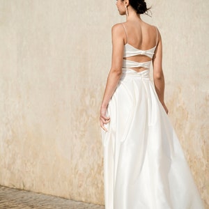 May include: A white satin wedding dress with a low back and a bow detail. The dress has a fitted bodice and a flowing skirt.