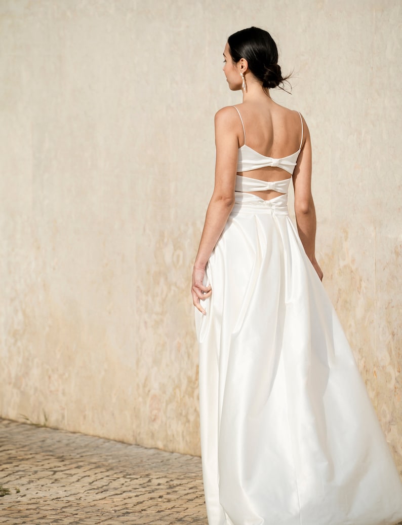 May include: A white satin wedding dress with a low back and a bow detail. The dress has a fitted bodice and a flowing skirt.