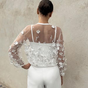 May include: White sheer top with floral embroidery, worn over a white vest top and white trousers.