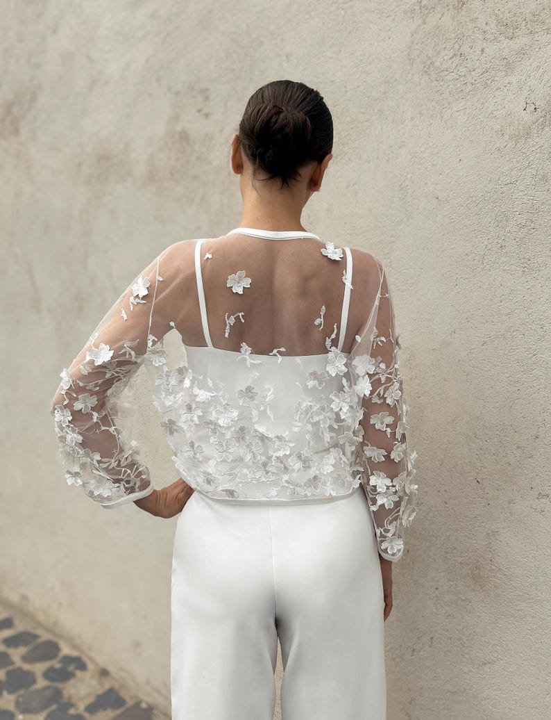 May include: White sheer top with floral embroidery, worn over a white vest top and white trousers.