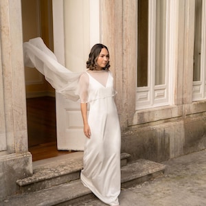 May include: A white, floor-length wedding dress with a sheer overlay and short sleeves. The dress has a V-neckline and a flowing skirt. A sheer, flowing fabric extends from the shoulders. The dress is worn outdoors near a stone building.
