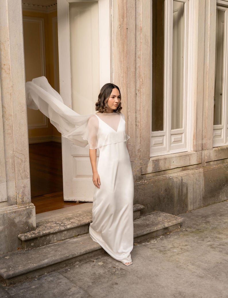 May include: A white, floor-length wedding dress with a sheer overlay and short sleeves. The dress has a V-neckline and a flowing skirt. A sheer, flowing fabric extends from the shoulders. The dress is worn outdoors near a stone building.