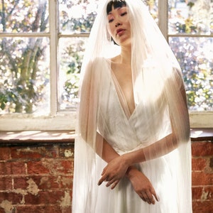 Puede incluir: Una mujer con un vestido de novia blanco y un velo transparente. Est&aacute; de pie frente a una ventana con luz natural entrando.