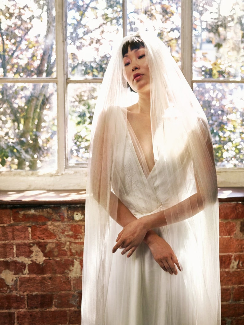 Puede incluir: Una mujer con un vestido de novia blanco y un velo transparente. Est&aacute; de pie frente a una ventana con luz natural entrando.