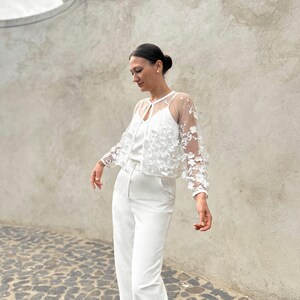 May include: A white lace top with a sheer overlay and floral embellishments. The top is paired with white wide-leg trousers.