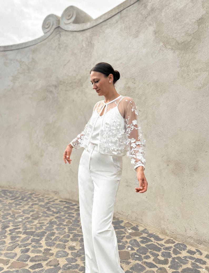 May include: A white lace top with a sheer overlay and floral embellishments. The top is paired with white wide-leg trousers.