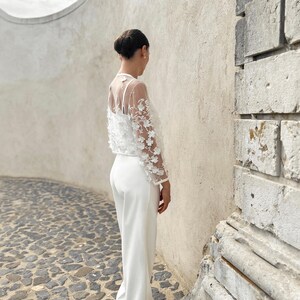 May include: A white trouser suit with a sheer lace top featuring floral embellishments. The suit is worn by a person standing against a white wall.