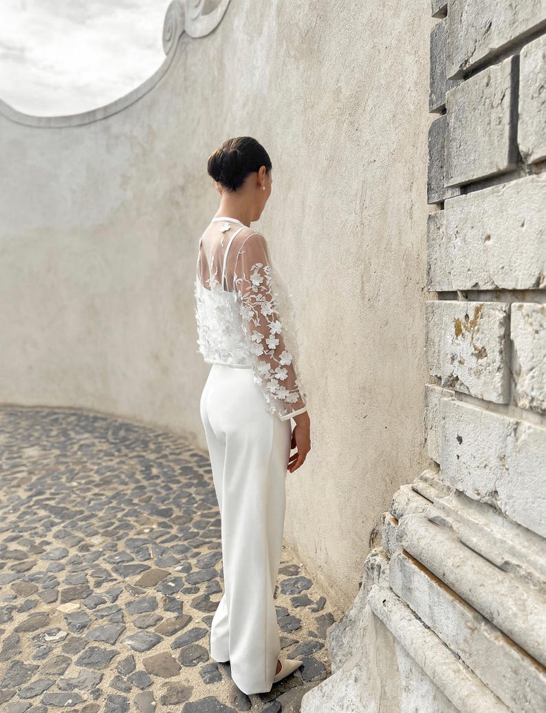 May include: A white trouser suit with a sheer lace top featuring floral embellishments. The suit is worn by a person standing against a white wall.