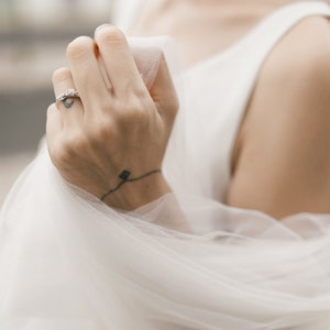 Puede incluir: Primer plano de una mano con un delicado anillo de diamantes. La mano sostiene un velo blanco transparente, creando una imagen suave y rom&aacute;ntica.