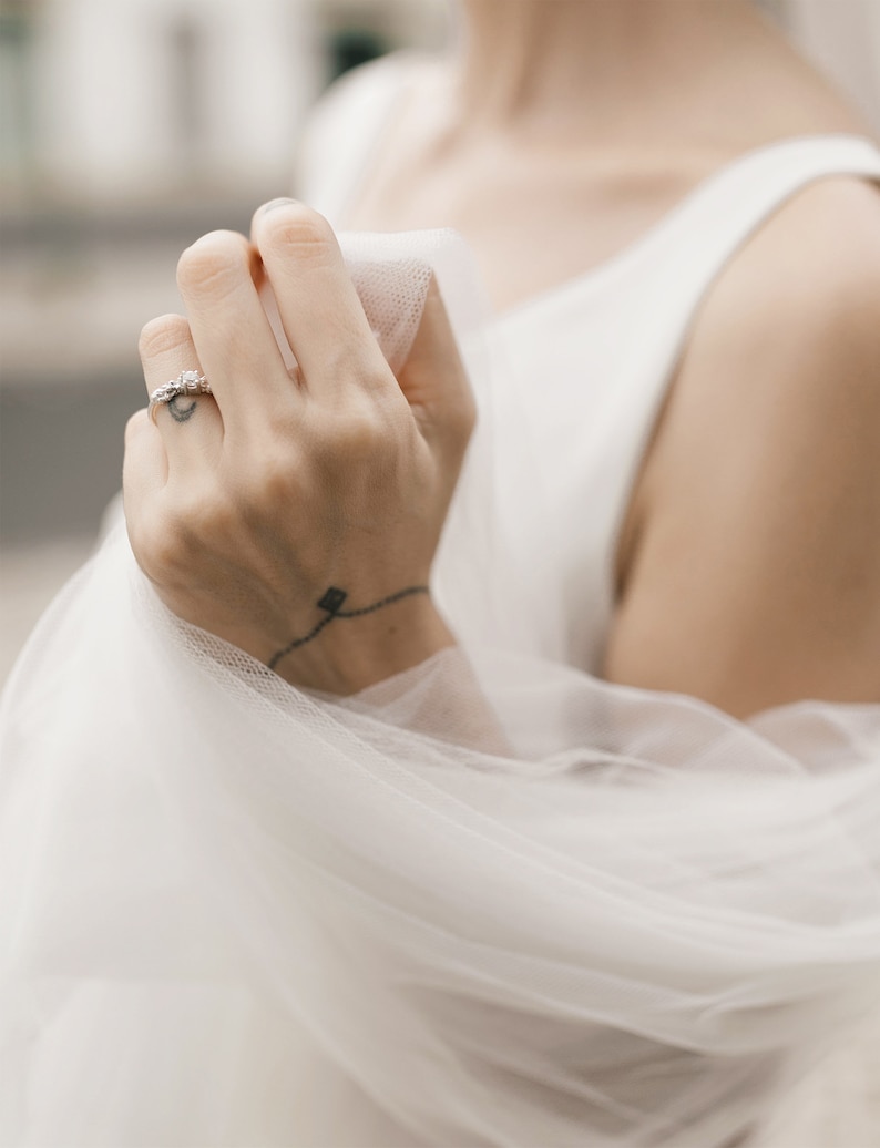 Puede incluir: Primer plano de una mano con un delicado anillo de diamantes. La mano sostiene un velo blanco transparente, creando una imagen suave y rom&aacute;ntica.