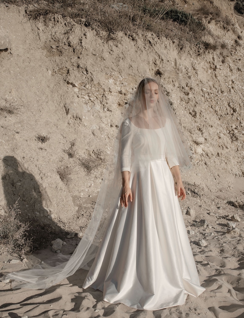 May include: A white wedding dress with a long veil. The dress has a fitted bodice and a flowing skirt. The veil is made of tulle and has a delicate lace trim.