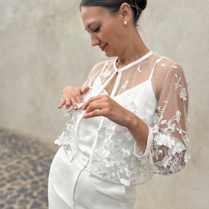May include: A white lace bolero with a floral design. The bolero has a tie closure at the neck and sheer sleeves.
